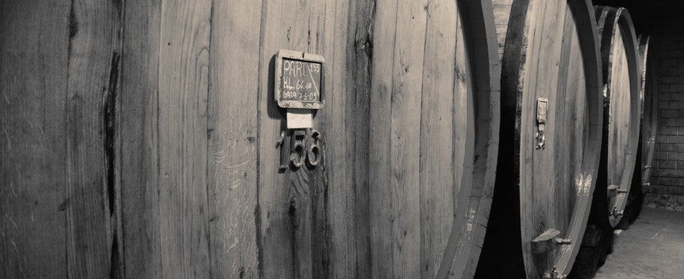 A row of large wooden barrels used for aging wine. Each barrel is marked with a small sign indicating details such as the batch number and date. The scene is set in a dimly lit cellar, emphasizing the rustic and traditional atmosphere of winemaking.
